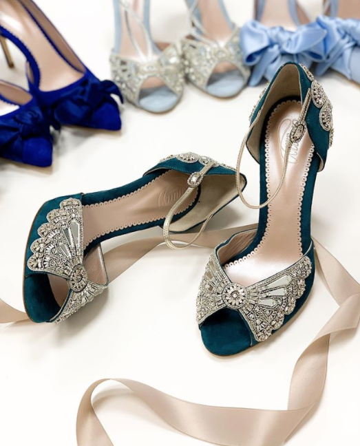 Teal suede heels with intricate silver embellishments, featuring an open toe and elegant ankle straps. The shoes are positioned on a white surface, surrounded by other colorful heels in the background, including blue and light blue options. The image is taken from a top-down angle, showcasing the detailed design of the heels. image