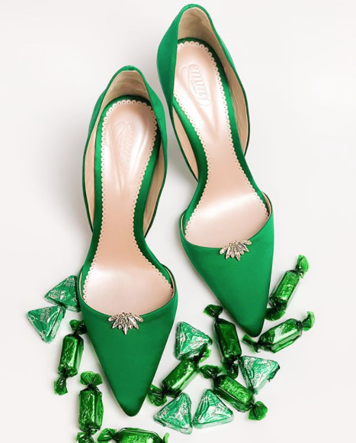 Green pointed-toe heels made of suede, featuring a decorative embellishment on the front. The shoes are positioned on a light background surrounded by green candy wrappers, showcasing a top-down view. image