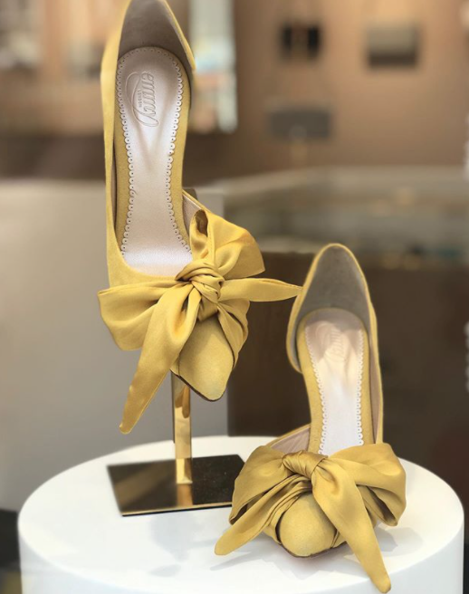 Yellow heels with a large bow detail at the front, displayed on a sleek gold stand. The background features a softly blurred boutique setting, enhancing the elegant presentation. The heels appear to have a mid-height heel. image