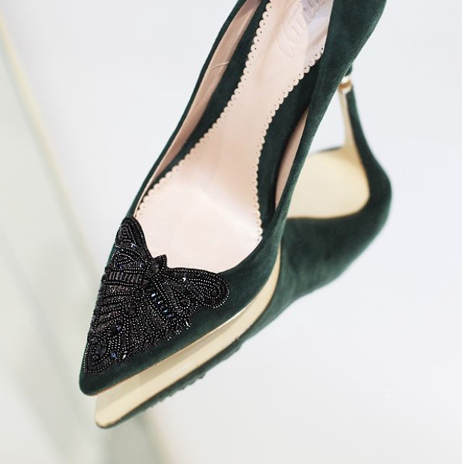 Green suede heels with a decorative black beaded butterfly design on the toe. The shoe is shown from a side angle, highlighting the elegant silhouette and the cut-out detail on the sides. The background is softly blurred, emphasizing the shoe's features. image