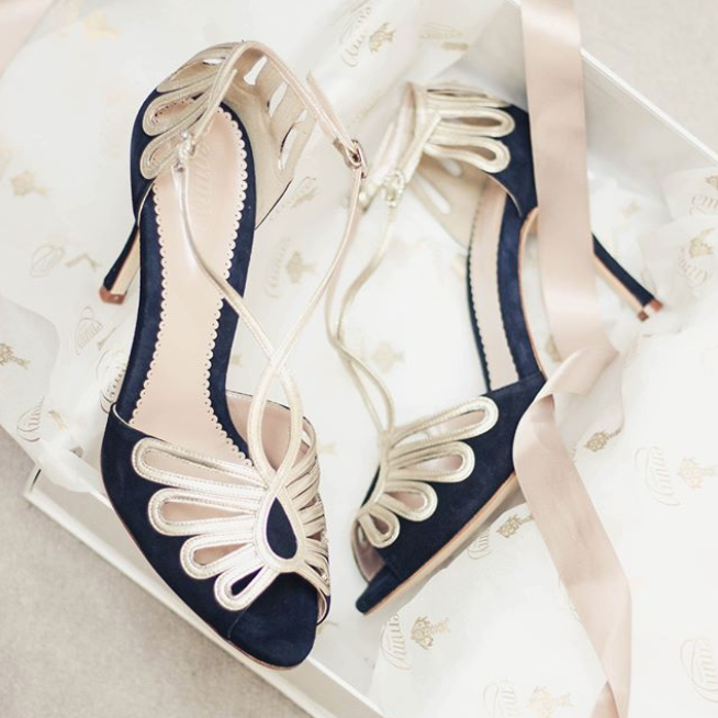 Elegant navy blue heels with a delicate gold strap design, displayed in a white box lined with tissue paper. The shoes feature a high heel and are made of a combination of suede and leather. The image is taken from a top-down angle, showcasing the intricate detailing of the straps. image