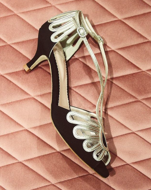 Elegant black heels with decorative gold accents, set against a soft pink quilted background. The shoes feature a mid-height heel and a delicate ankle strap, showcasing a stylish design. The image captures the shoes from a top-down angle, emphasizing their intricate detailing. image