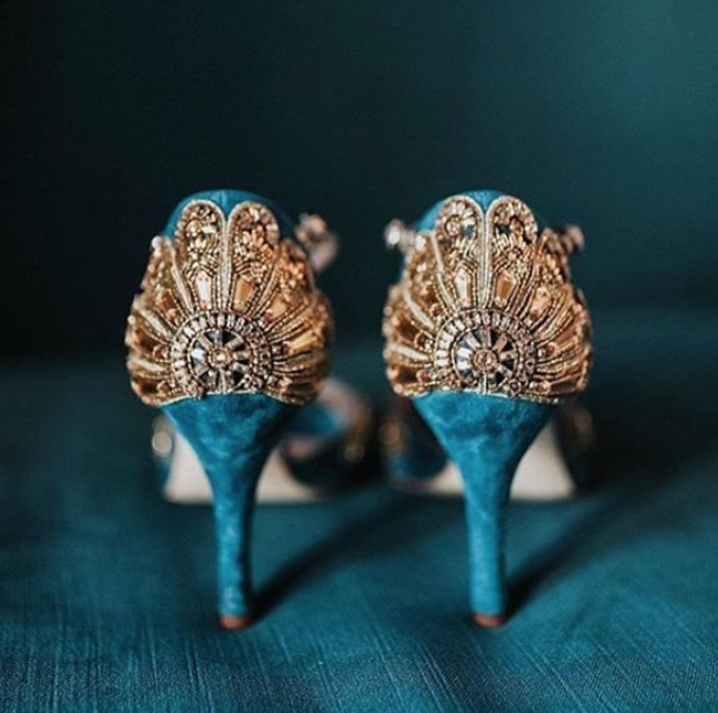 Elegant blue suede heels featuring intricate gold embellishments at the back. The image is taken from a close angle, showcasing the detailed design against a rich teal background. The heels are high, adding a touch of glamour to the overall look. image