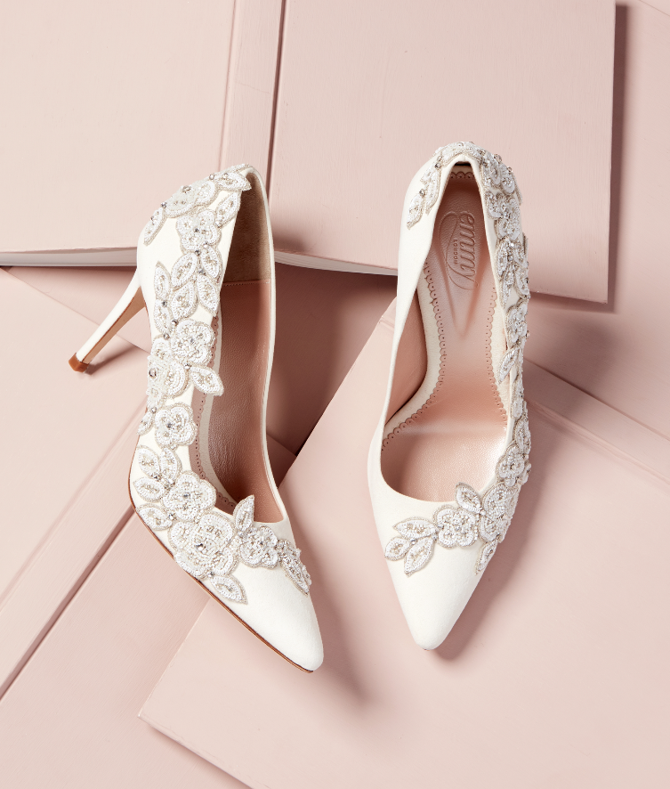 Elegant white heels adorned with intricate floral embellishments, set against a soft pink background. The shoes feature a pointed toe and a slender stiletto heel, showcasing a sophisticated design. image